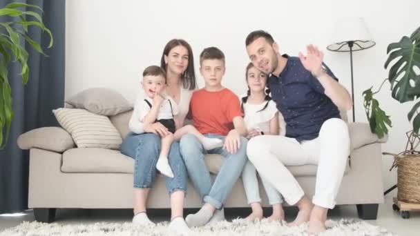 Onnellinen suuri eurooppalainen perhe rentoutuu kotona sohvalla, he heiluttavat iloisesti käsiään kameralle. Perheen ja onnellisuuden käsite - Materiaali, video
