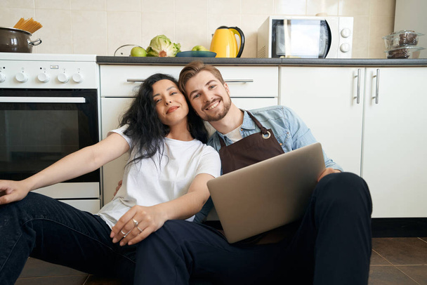 Romantic guy with laptop hugging pleased lady seated beside him on floor - Fotó, kép