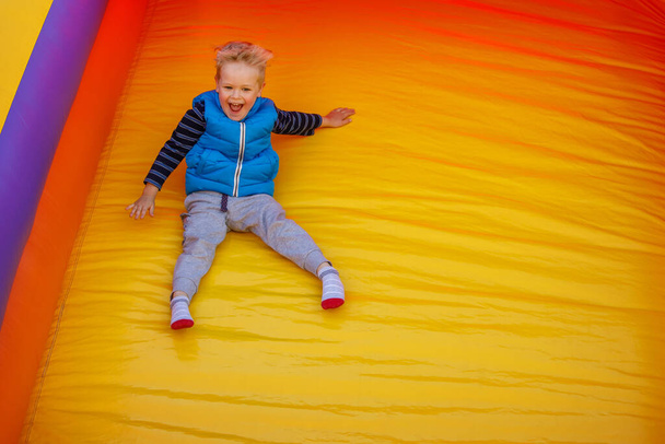 Chlapec skáče ze skluzavky na žlutooranžovém nafukovacím skákacím hradě. Na obrázku je volné místo pro text. - Fotografie, Obrázek