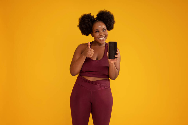 Smiling, happy, young girl with afro hairstyle posing on yellow studio background in sporty clothes, holding mobile phone in one hand showing new fitness app. Okay sign. Sale concept. Copy space.  - Foto, imagen