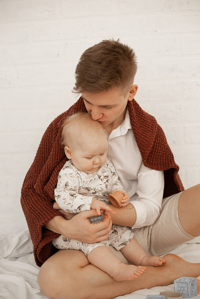 Muotokuva nuori komea isä isä peitetty ruudullinen tilalla, suudella pieni pullea vauva, leikkii moderni pastelli silikoni kulhot erikokoisia ja kuutiot eri kuvioita valkoisella sängyllä.  - Valokuva, kuva
