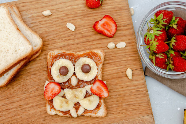 Funny owl face sandwich toast bread with peanut butter, banana, strawberry on wooden board. Cute kids childrens baby's sweet dessert, healthy breakfast,lunch, food art on gray background,top view. - Fotoğraf, Görsel