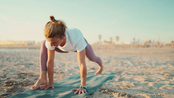 Deportiva rubia ejercitando yoga sobre colchoneta de fitness junto al mar. Atractiva chica yogui vestida con ropa deportiva haciendo práctica matutina en la playa - Foto, Imagen