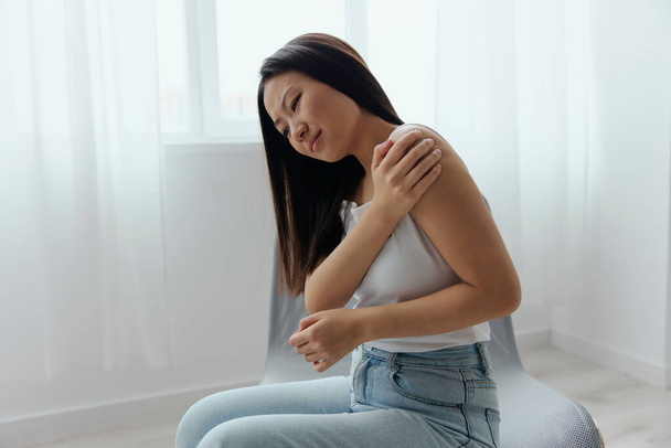 Getroffen door schouderontwrichting fracturen pijn na het optillen van gewichten in de verkeerde positie gebruinde mooie jonge Aziatische vrouw aanraken pijnlijke schouder thuis huiskamer. Cool aanbod. - Foto, afbeelding