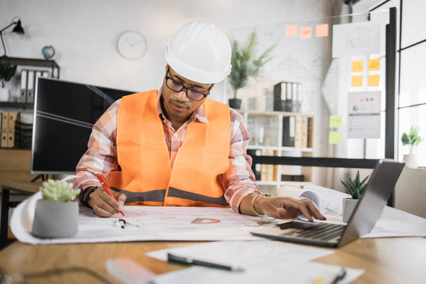 Portrait of african american man wearing uniform and white hard hat at work. Confident contractor sitting at the desk, holding pen and working on laptop with sketches of building object. - Φωτογραφία, εικόνα