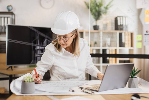Mature architect woman or female builder holding pen while writing and drawing architectural project using laptop computer, sitting on the desk at workplace in modern office. - Photo, Image
