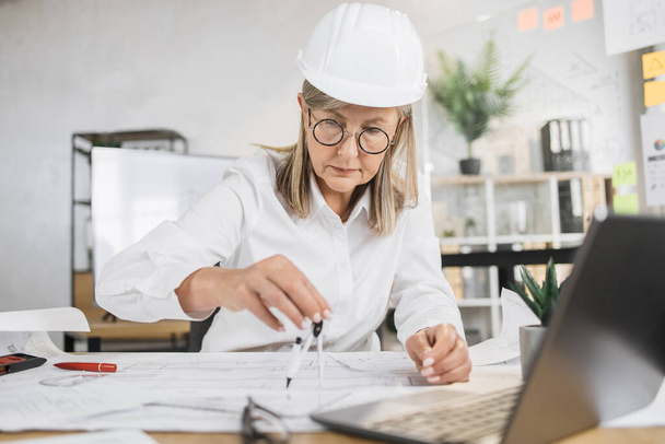 Competent mature female engineer, designer or architect leading working conference at office center. Attractive young senior woman wearing protective helmet working with compass, laptop and blueprint. - Фото, изображение