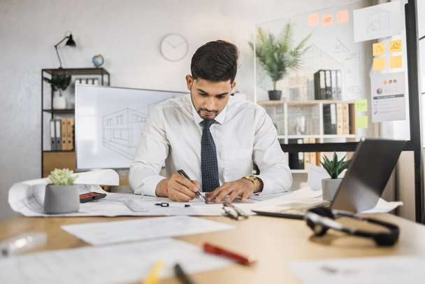 Competent arhitect indian male sitting on desk indoors using pen and ruler for project drawings. Positiv hindu bearded young man wearing white shirt and black tie. - Foto, Imagem