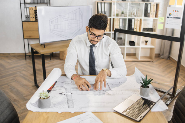 Qualified arab architect in white shirt, tie and glasses checking construction project with blueprint and laptop sitting on table. Bearded attractive muslim man controlling working process. - Φωτογραφία, εικόνα