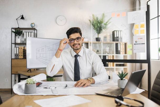 Young architect arab male in white shirt and tie sitting at table with blueprints and modern devices. Focused man worker creating new building project sitting in modern office. - Foto, Imagen