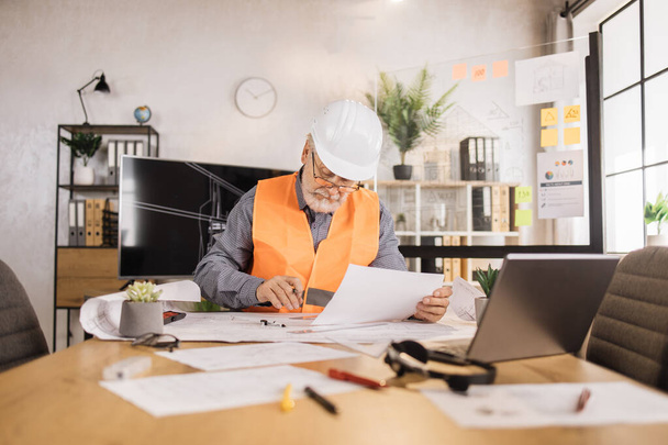 Senior builder, engineer in reflective vest uniform and hard hat on desk in office on background of large TV screen with , browsing in laptop and looking through papers or sketches. - Foto, afbeelding