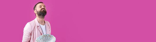 Tyytyväisen nuoren liikemiehen muotokuva, jolla on parta pukeutuneena vaaleanpunaiseen takkiin, näyttää meille dollarin seteleitä punaisen studion taustaa vasten. Maku, rahan haju. - Valokuva, kuva
