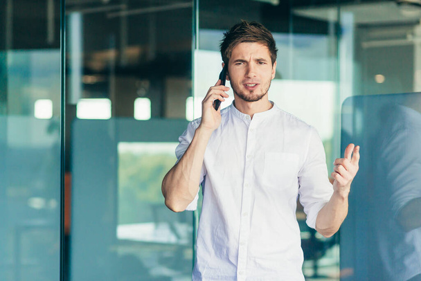 Беспокойный молодой бизнесмен разговаривает по мобильному телефону возле офиса. Размахивая руками, недовольный - Фото, изображение