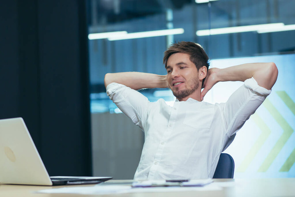 Young handsome man freelancer, businessman, manager in the office at the table resting, doing work during a break, throwing his hands behind his head, smiling - Foto, Bild