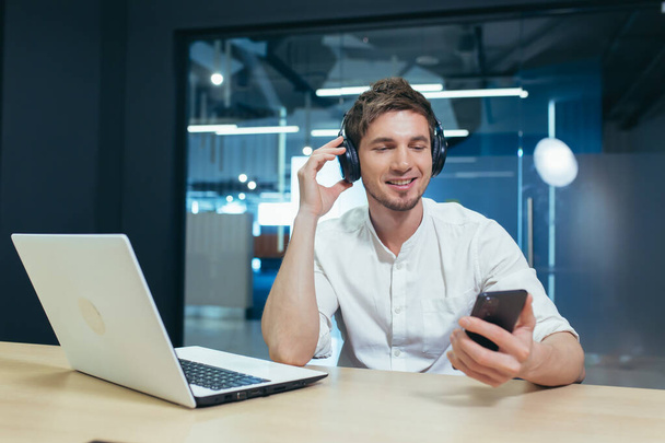 A young man listens to podcasts in headphones, holds a mobile phone in his hands, talks on video communication. Sitting at work in the office with a laptop - 写真・画像