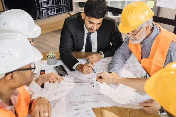 Qualified architects, engineers and designers having business meeting at office. Multicultural colleagues in suits and helmets sitting at table and looking at blueprints. Construction concept. - 写真・画像