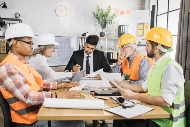 Multiracial men and women in formal outfit and helmets sitting at table with lots of blueprints during important strategic session. Business people working on common construction project. - 写真・画像