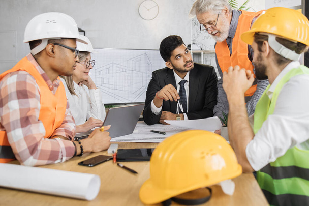 Multicultural partners working with drawings inspection during conference at modern office. Business colleagues wearing suits, uniform and protective helmet. Construction concept. - Foto, Imagem