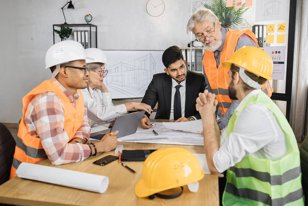 Multicultural partners working with drawings inspection during conference at modern office. Business colleagues wearing suits, uniform and protective helmet. Construction concept. - 写真・画像