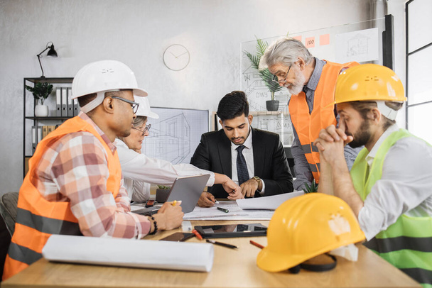 Group of five multiracial people in suits and helmets brainstorming for common project of new building. Construction team working with blueprints and modern devices at office. - Photo, image