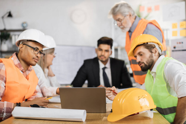 Group of coworkers in suits and helmets having business meeting at office. Responsible engineers focusing on common project of new building. Construction industry. - Foto, Imagen