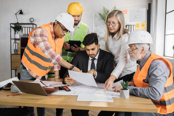 Groep architecten en ingenieurs zitten op kantoor en werken aan een gemeenschappelijk project. Indiase baas man in business suit zit aan tafel en bespreekt bouwstrategie met collega 's en partners. - Foto, afbeelding