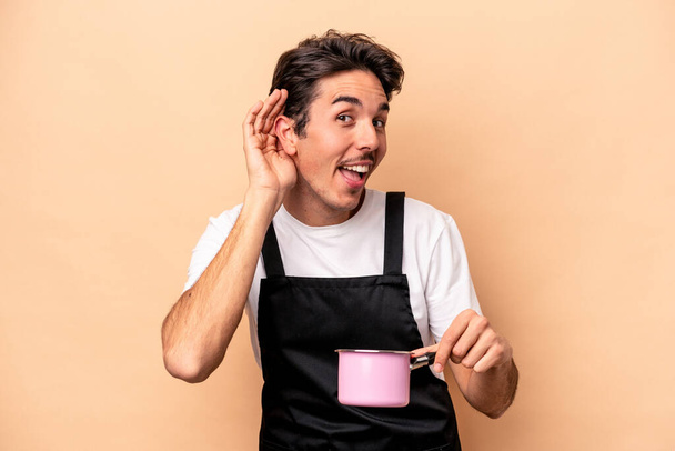 Young caucasian man holding a saucepan isolated on beige background trying to listening a gossip. - Photo, Image
