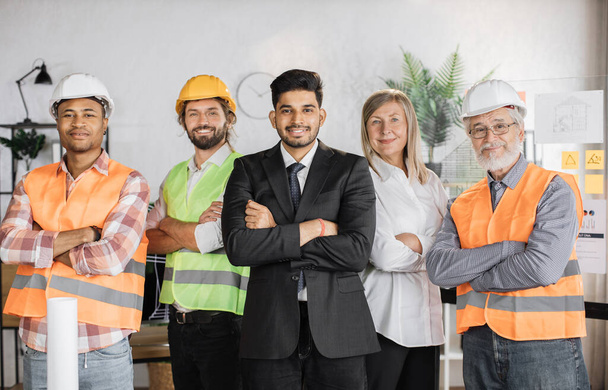 Front view of one senior woman and four men engineers, builders, architects looking at camera while standing on light modern office. People of different age, gender and race. - Foto, Imagem