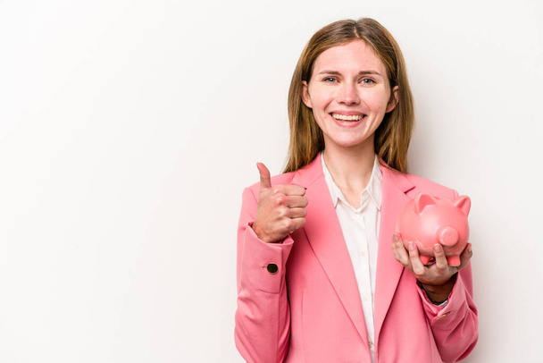 Young business English woman holding a piggybank isolated on white background smiling and raising thumb up - Foto, imagen