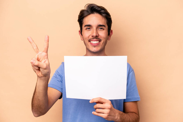 Young caucasian man holding a placard isolated on beige background showing number two with fingers. - Photo, Image