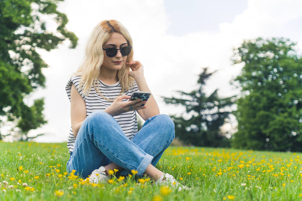 Pełne ujęcie blondynki europejskiej na trawie przewijającej portale społecznościowe. Wysokiej jakości zdjęcie - Zdjęcie, obraz