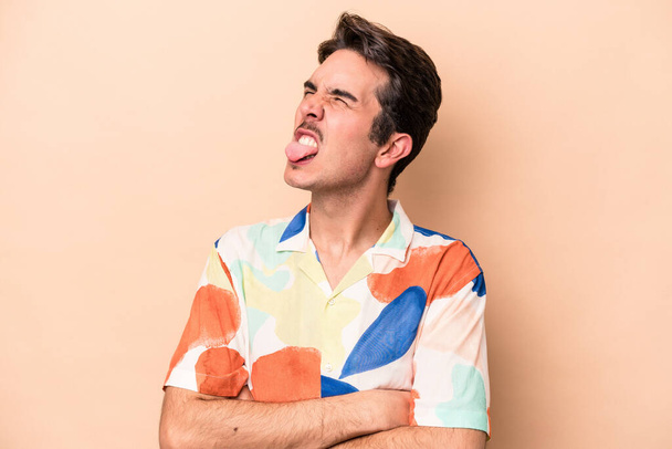 Young caucasian man isolated on beige background funny and friendly sticking out tongue. - Photo, Image