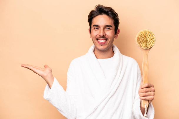 Young caucasian man wearing a bathrobe holding a back scratcher isolated on beige background showing a copy space on a palm and holding another hand on waist. - Photo, Image
