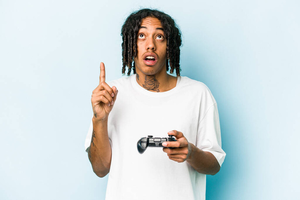 Young African American man playing with a video game controller isolated on blue background pointing upside with opened mouth. - Zdjęcie, obraz