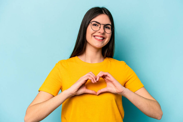 Young caucasian woman isolated on blue background smiling and showing a heart shape with hands. - Fotografie, Obrázek