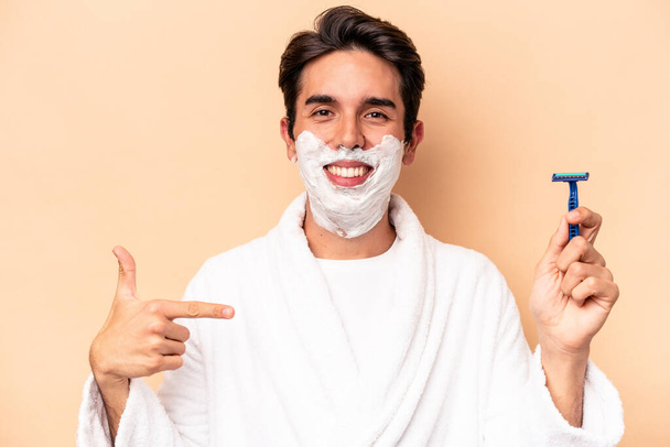 Young caucasian man shaving his beard isolated on beige background person pointing by hand to a shirt copy space, proud and confident - Фото, зображення