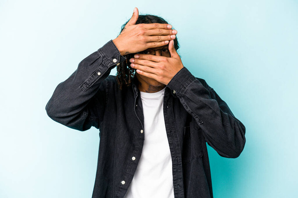 Jovem afro-americano isolado no fundo azul pestanejar para a câmera através dos dedos, rosto de cobertura envergonhado. - Foto, Imagem