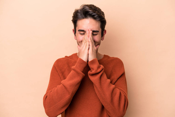 Νεαρός καυκάσιος άνδρας απομονωμένος σε μπεζ φόντο κρατώντας τα χέρια στο προσεύχομαι κοντά στο στόμα, αισθάνεται αυτοπεποίθηση. - Φωτογραφία, εικόνα