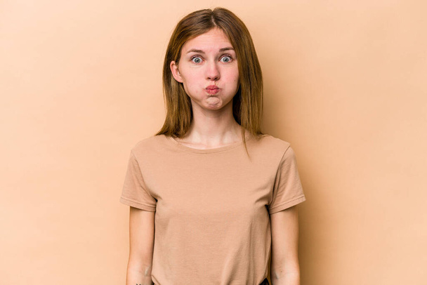 Young English woman isolated on beige background blows cheeks, has tired expression. Facial expression concept. - Photo, image