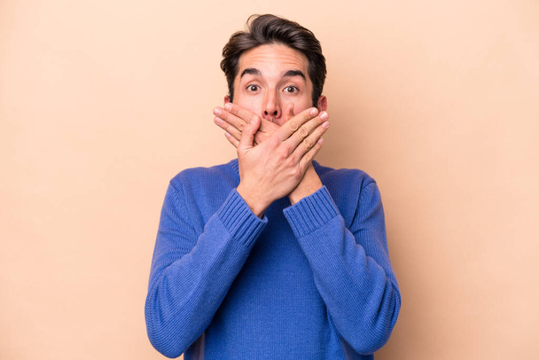 Young caucasian man isolated on beige background shocked covering mouth with hands. - Foto, afbeelding
