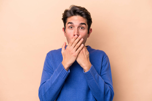 Young caucasian man isolated on beige background shocked, covering mouth with hands, anxious to discover something new. - Foto, Imagen