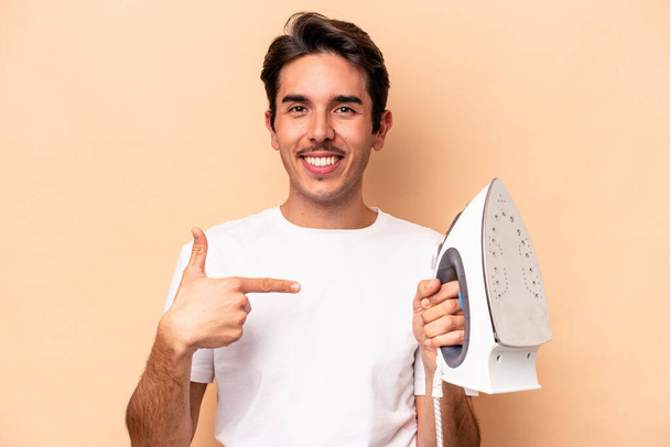 Young caucasian man holding an iron isolated on beige background person pointing by hand to a shirt copy space, proud and confident - Valokuva, kuva