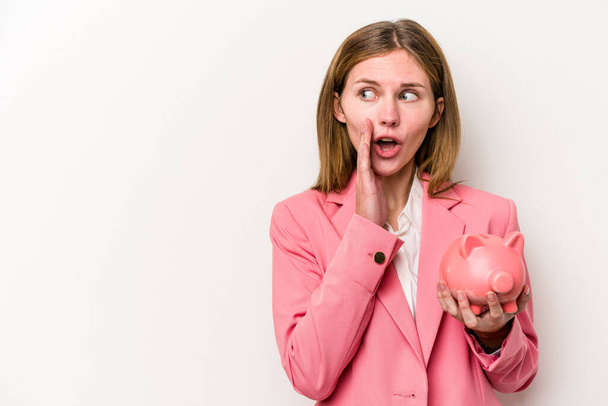 Young business English woman holding a piggybank isolated on white background is saying a secret hot braking news and looking aside - Photo, Image