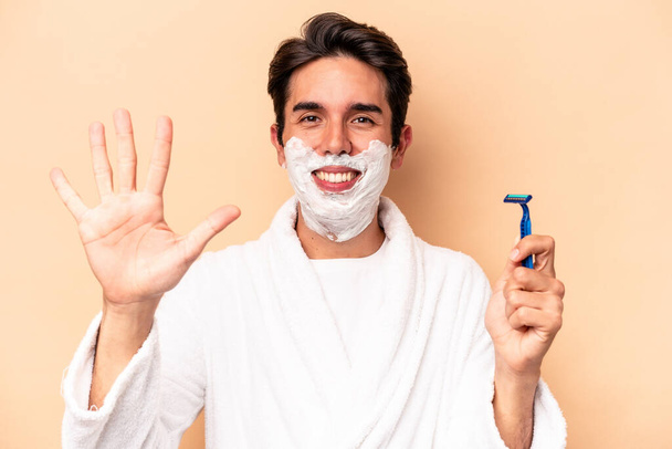 Jovem caucasiano barbear sua barba isolada no fundo bege sorrindo alegre mostrando número cinco com os dedos. - Foto, Imagem
