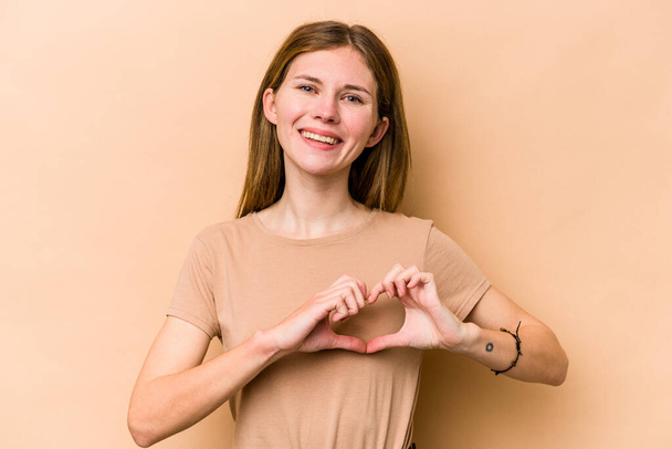 Young English woman isolated on beige background smiling and showing a heart shape with hands. - Fotografie, Obrázek