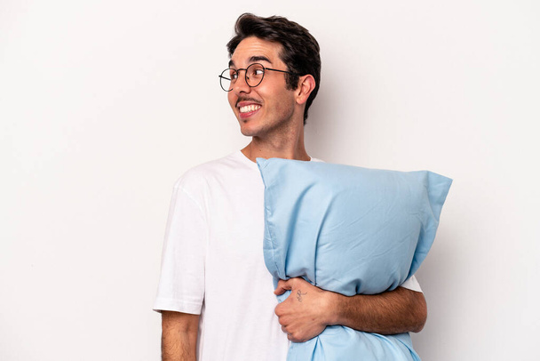Jovem caucasiano vestindo pijama segurando um travesseiro isolado no fundo branco olha para o lado sorridente, alegre e agradável. - Foto, Imagem