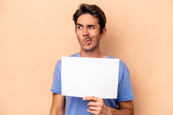 Young caucasian man holding a placard isolated on beige background confused, feels doubtful and unsure. - 写真・画像