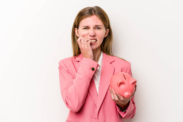 Young business English woman holding a piggybank isolated on white background biting fingernails, nervous and very anxious. - Photo, Image