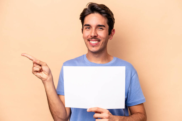 Młody biały mężczyzna trzymający tabliczkę odizolowaną na beżowym tle uśmiechający się i odsuwający na bok, pokazujący coś w pustej przestrzeni. - Zdjęcie, obraz