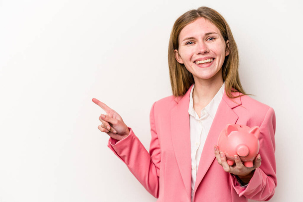 Young business English woman holding a piggybank isolated on white background smiling and pointing aside, showing something at blank space. - Fotografie, Obrázek